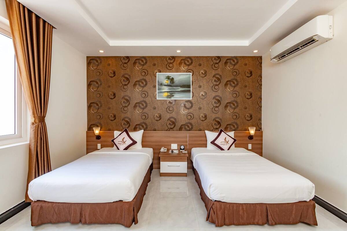 A hotel room with two beds, a nightstand, a wall-mounted air conditioner, and a painting of a landscape.
