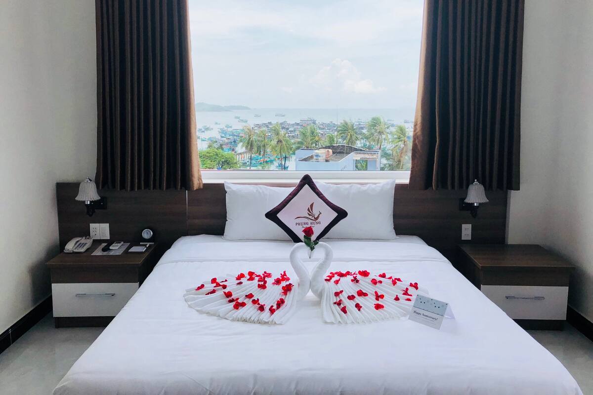 A hotel room with a bed, a nightstand, a lamp, and a view of the sea and palm trees.