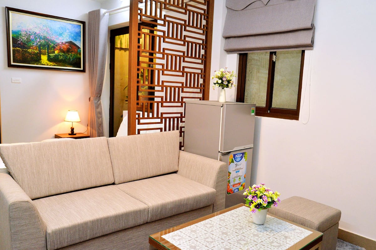Ein Wohnzimmer mit einer Couch, einem kleinen Tisch mit Blumenvase, einem Kühlschrank und einem dekorativen Raumteiler.