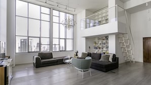 A spacious living room with large windows, a mezzanine level with bookshelves, and a staircase. - Deluxe Holiday Homes - East Heights 1 (Dubai)