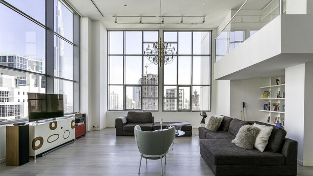 A modern living room with a large window, a grey sofa, a small armchair, and a TV on a wooden cabinet. - Deluxe Holiday Homes - East Heights 1 (Dubai)