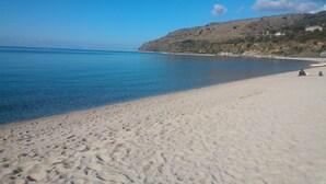 Une plage de sable fin bordée d’une eau calme, un ciel dégagé et un paysage vallonné en arrière-plan.