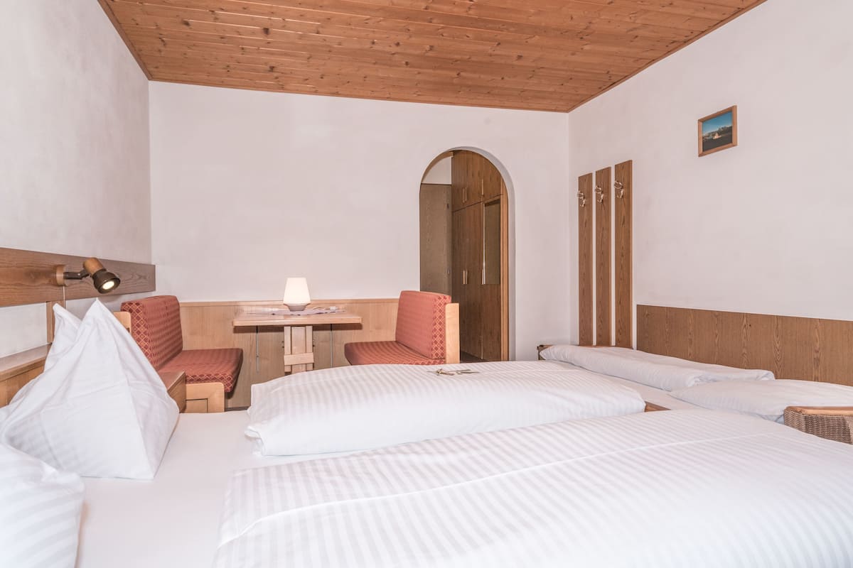 Ein Hotelzimmer mit Holzboden, einem Bett mit weißen Leinentüchern, einem kleinen Schreibtisch mit Lampe, einem roten Polsterstuhl und einem Holzschrank.