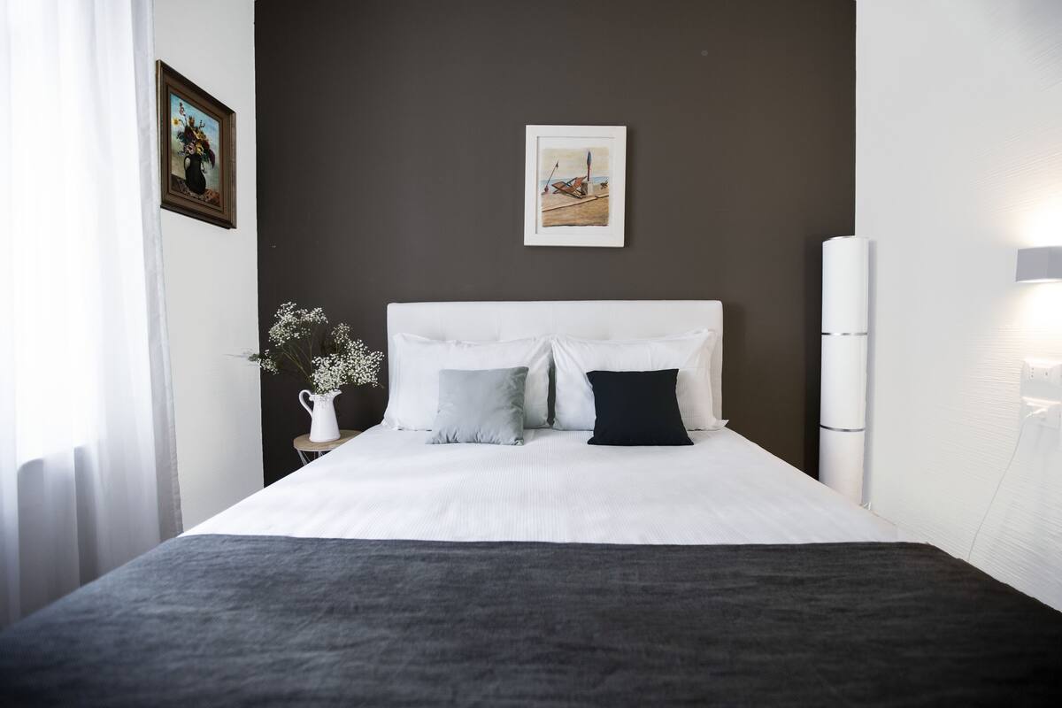 A neatly made bed with white and dark grey bedding, a white headboard, and a small white table beside it with a vase of flowers.