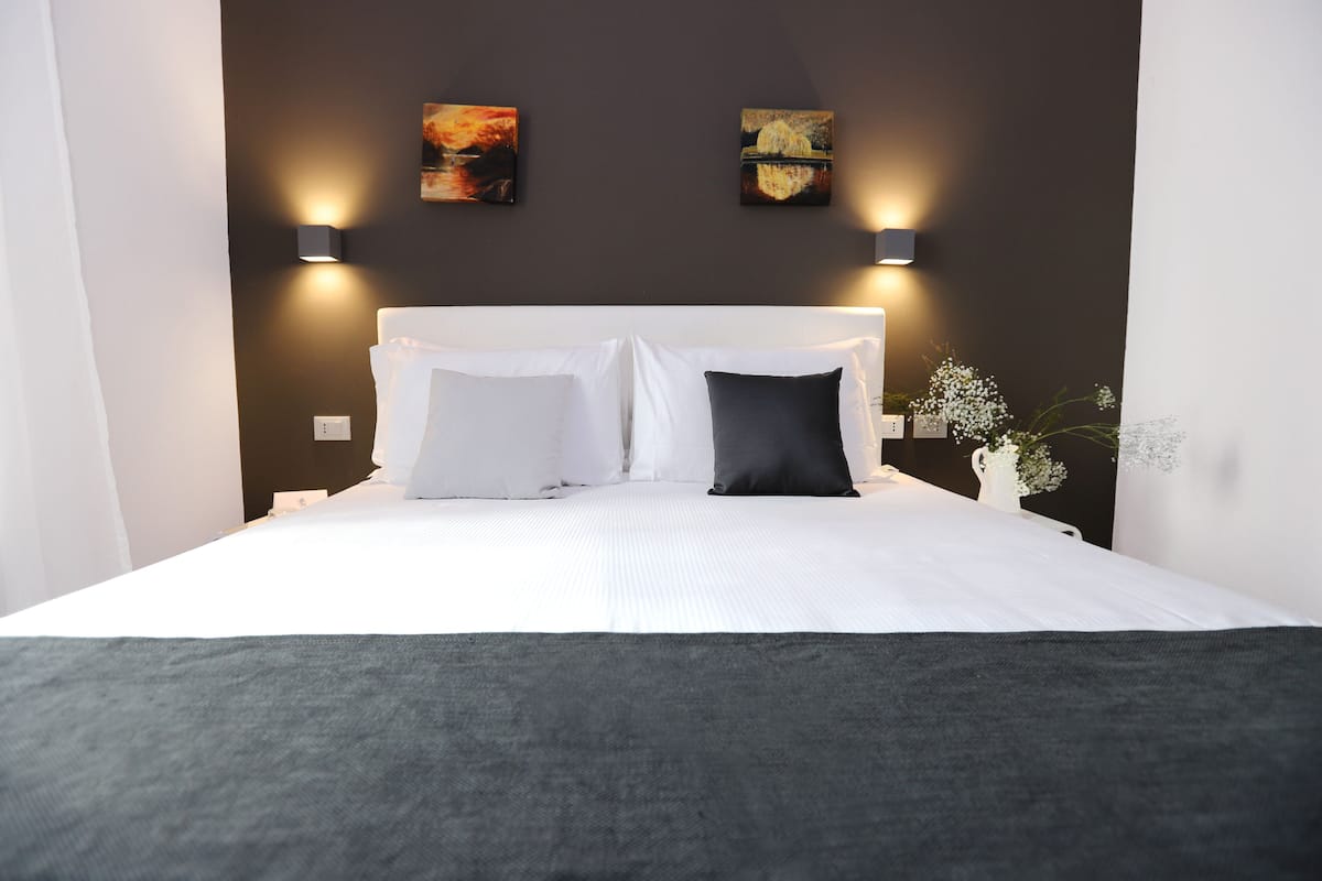 A neatly made bed with white and dark pillows, two wall-mounted light fixtures, and a small vase with white flowers on the right side.