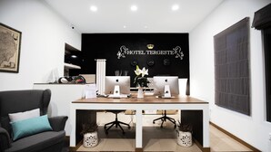 A modern hotel reception desk with two Apple iMac computers, a black wall with the hotel's name and lion logo, a grey sofa with teal cushions, and two potted plants. - Hotel Tergeste (Trieste)