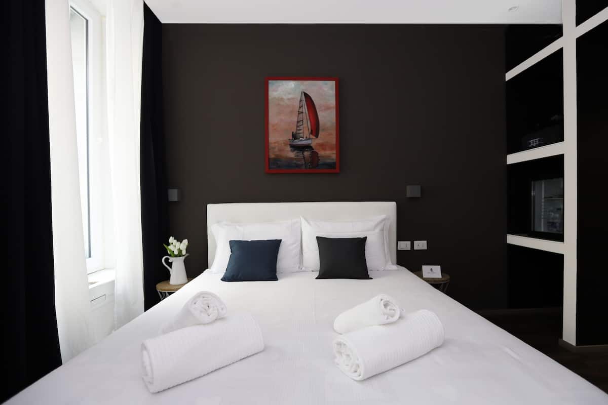 A hotel room with a large bed, white linens, a painting of a sailboat, and a small table with a vase of flowers.