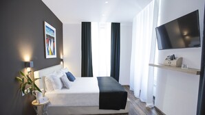 A modern hotel room with a large bed, a flat-screen TV mounted on the wall, a small side table with a lamp, and a large window with curtains. - Hotel Tergeste (Trieste)