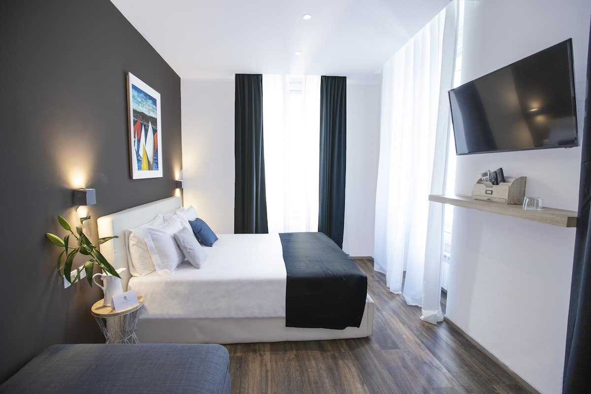 A modern hotel room with a large bed, a flat-screen TV mounted on the wall, a small side table with a lamp, and a large window with curtains.