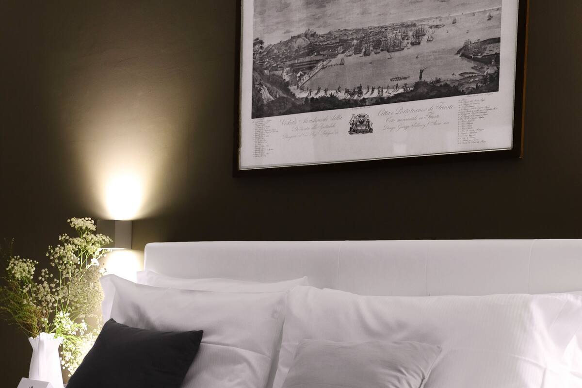 A neatly made bed with white linens and two pillows, a framed black and white photograph on the wall, and a bedside table with a vase of flowers.