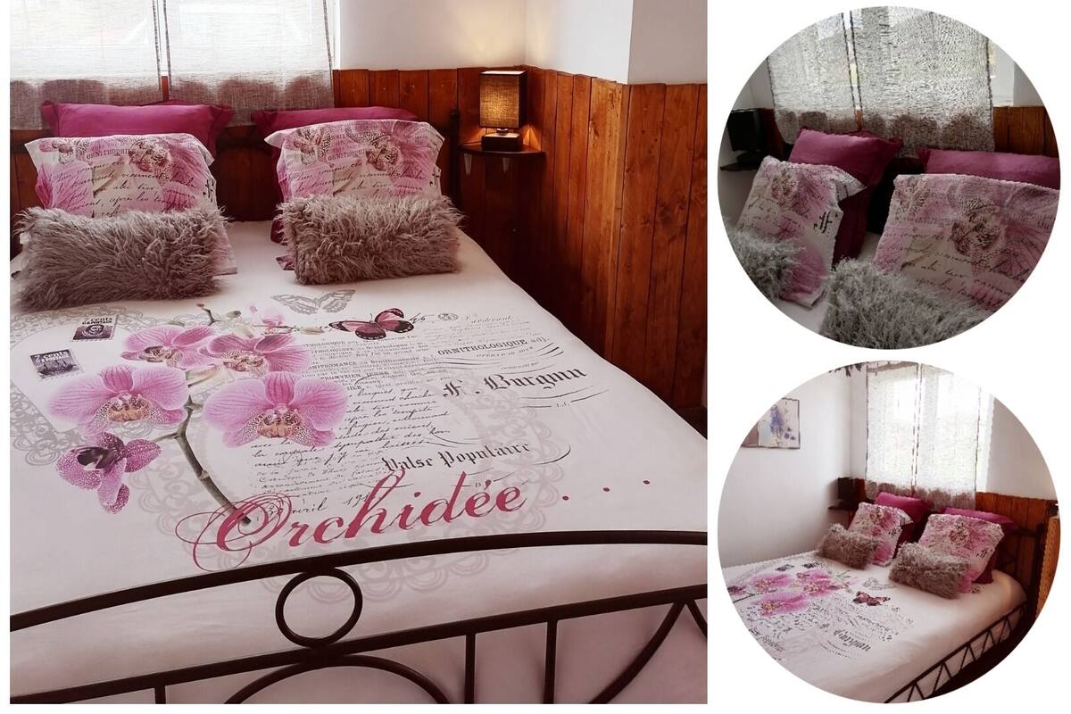 A bedroom with a bed covered in a floral-patterned bedspread, multiple fluffy pillows, and a nightstand with a lamp.