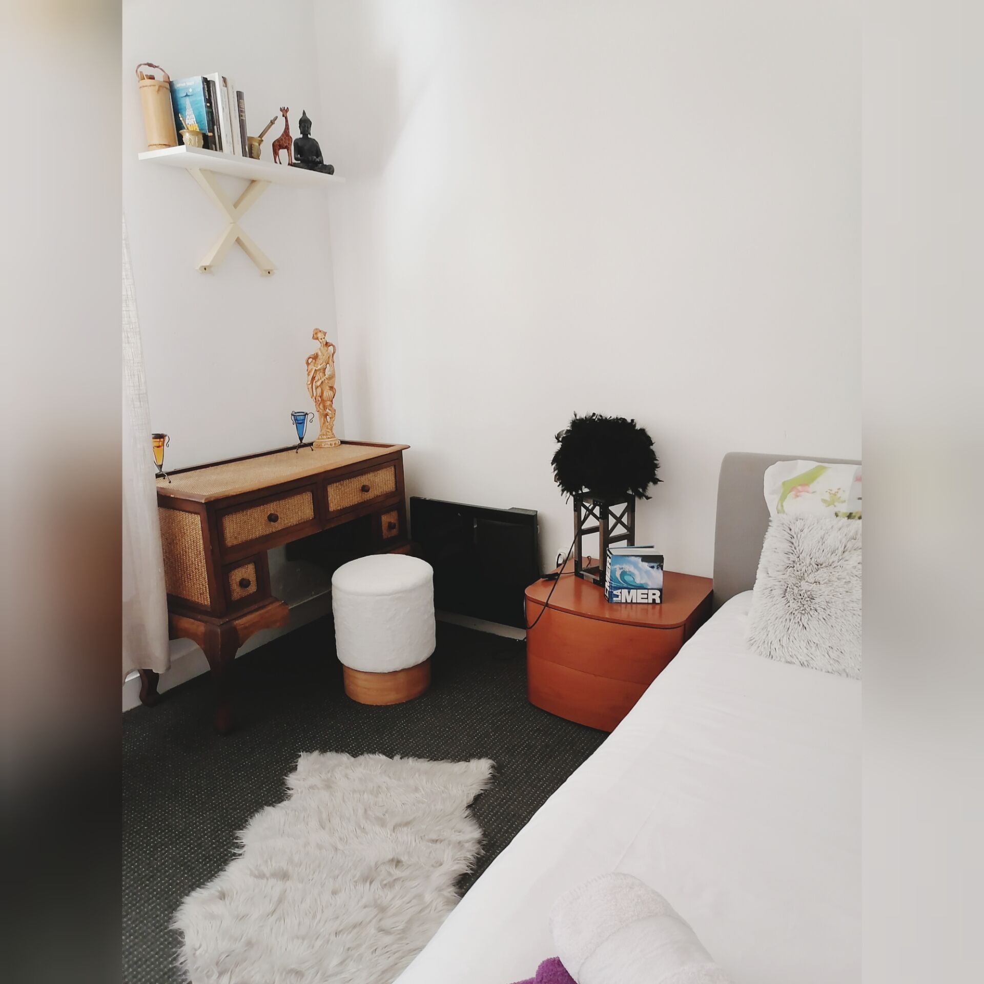 Een slaapkamer met een houten bureau, een witte poef, een bed met een wit en paars kussen, en een klein plankje met decoratieve objecten.