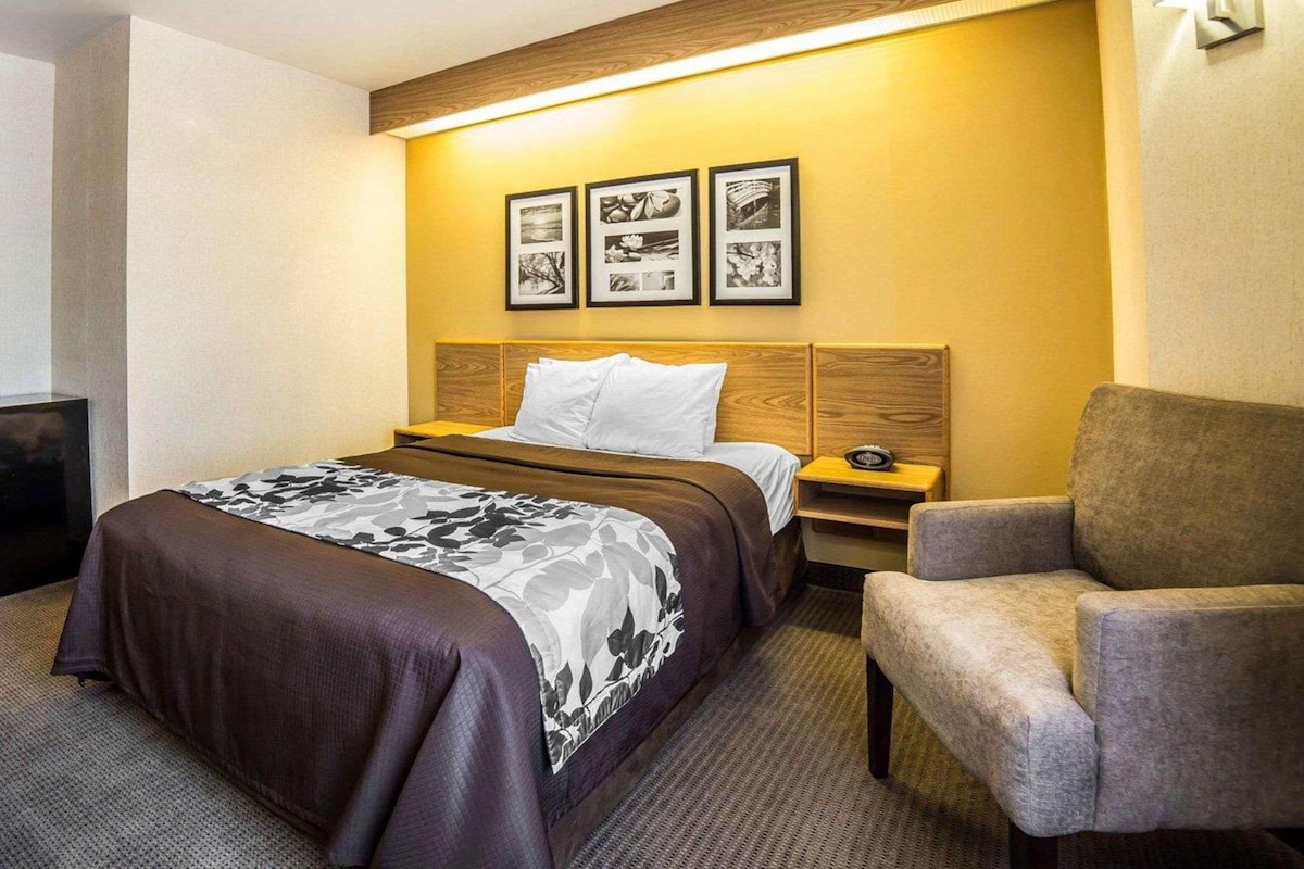 A hotel room with a bed, a chair, a nightstand, and a wall-mounted lamp.