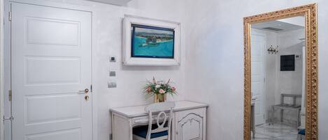 A hotel room with a white door, a desk with a chair, a framed picture on the wall, and a large mirror with a gold frame.