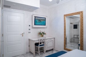 A hotel room with a white door, a desk with a chair, a framed picture on the wall, and a large mirror with a gold frame.