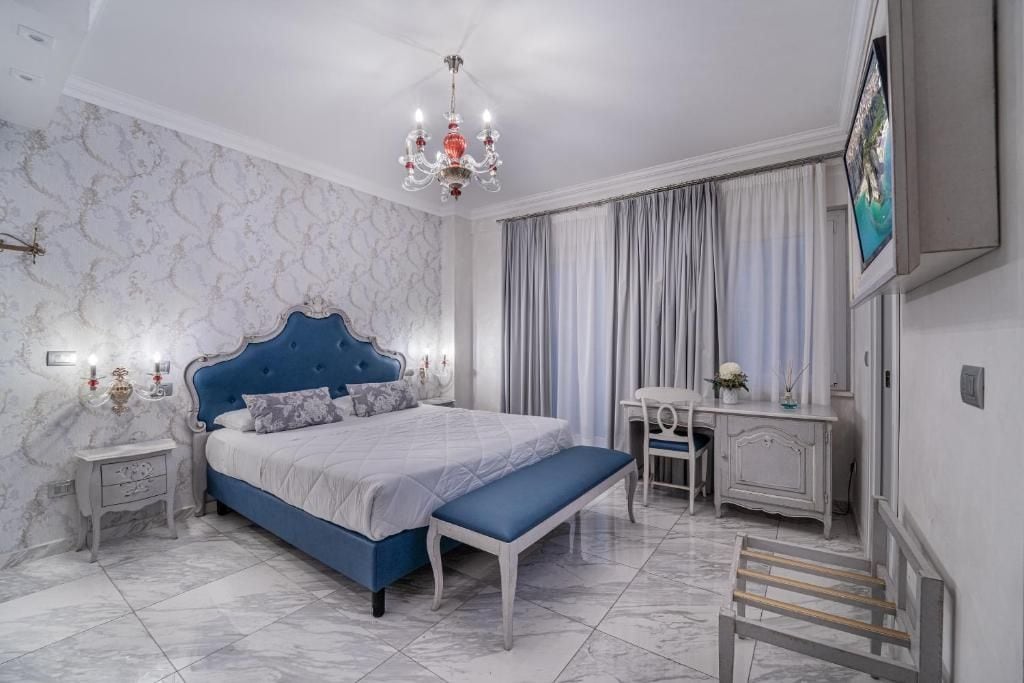 A bedroom with a large bed, a bench, a desk, a chair, a TV, and a chandelier.