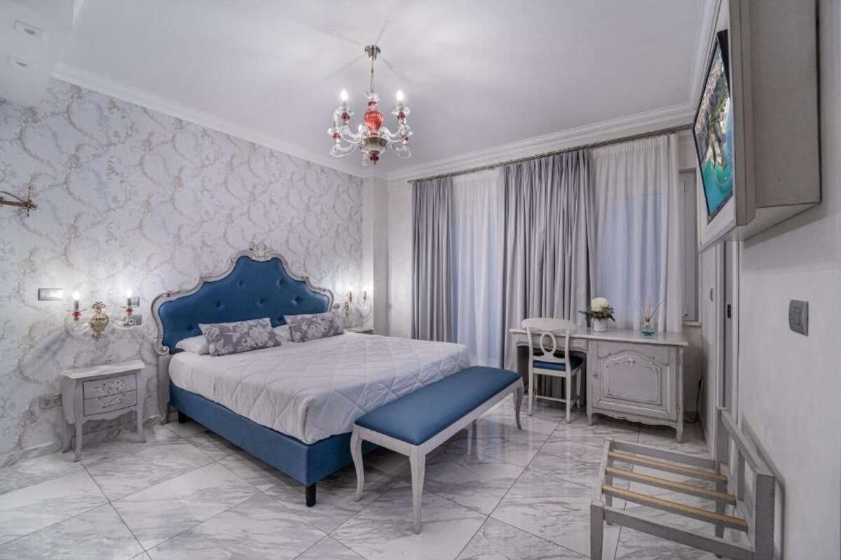 Ein Schlafzimmer mit einem großen Bett, einer Bank, einem Schreibtisch, einem Sessel, einem Fernseher und einer Kronleuchter.