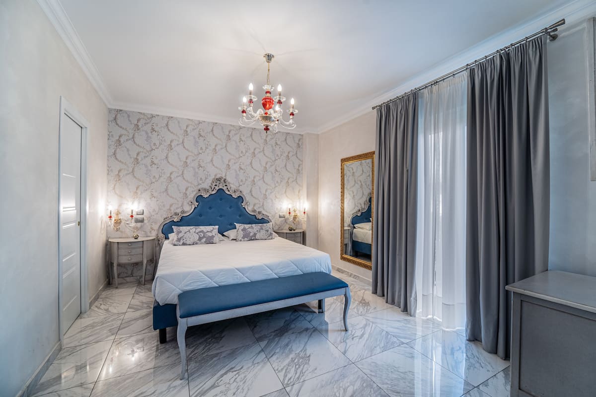 A bedroom with a marble floor, a large bed with a blue headboard, a chandelier, a mirror, and grey curtains.
