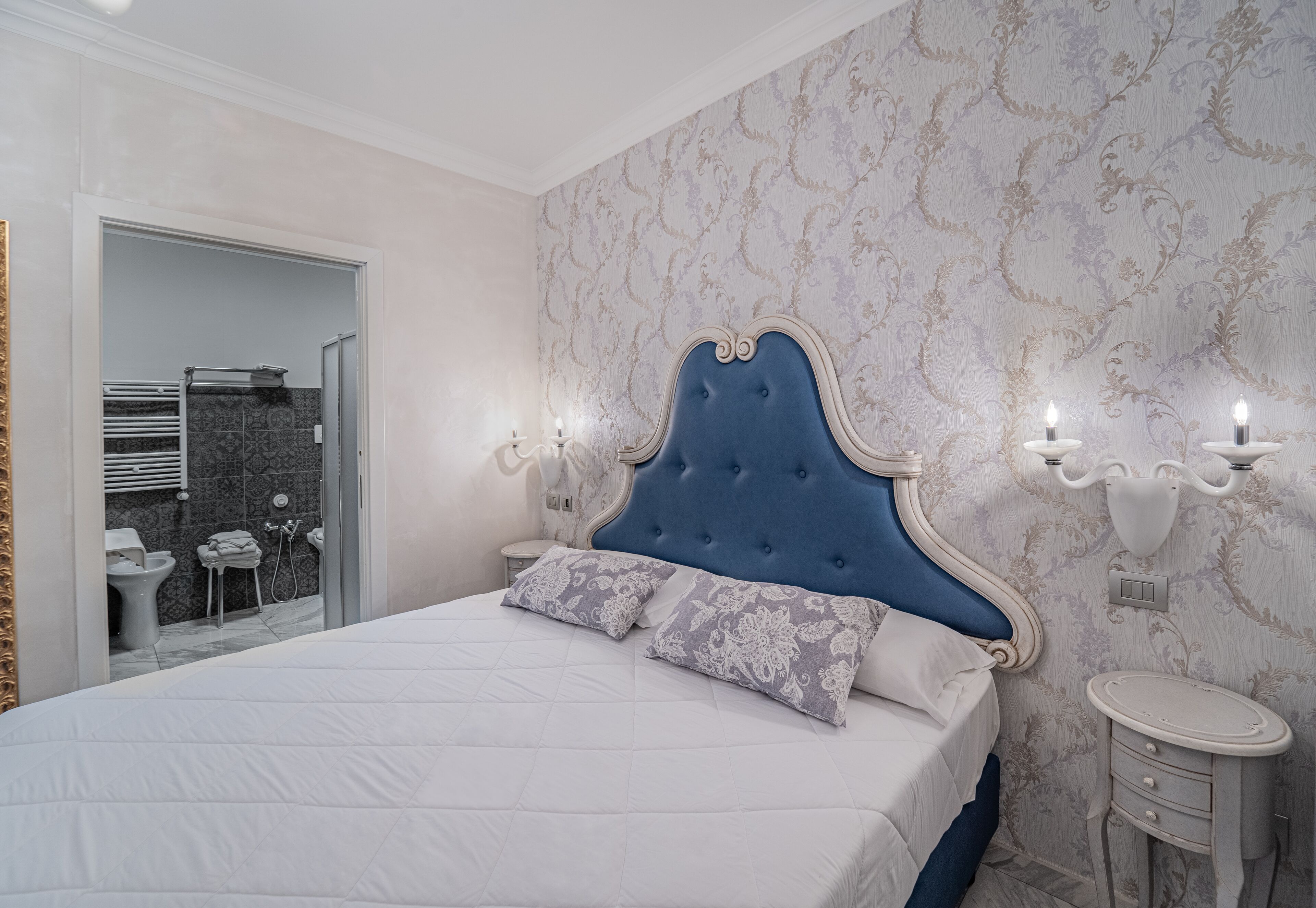 Un dormitorio con una cama grande, una mesita de noche, una lámpara de pared y un baño visible a través de una puerta abierta.