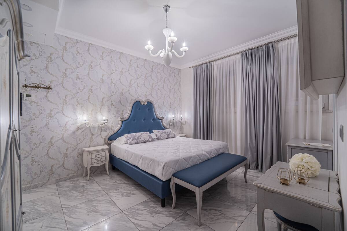 A bedroom with a blue bed, a white nightstand, a marble floor, a chandelier, and a large window with curtains.