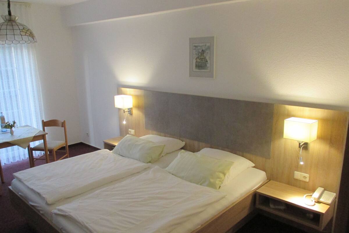 A hotel room with a double bed, two bedside tables with lamps, a small table with a chair, and a picture on the wall.