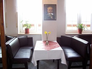 A dining area with two black leather booths, a white table with a red placemat, and potted plants on windowsills. - Hotel Reuterhof (Stavenhagen)