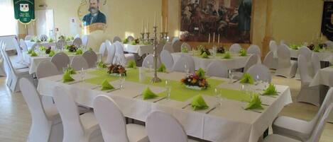 Une salle de banquet avec de longues tables dressées pour le repas, des chaises blanches et un grand tableau accroché au mur.