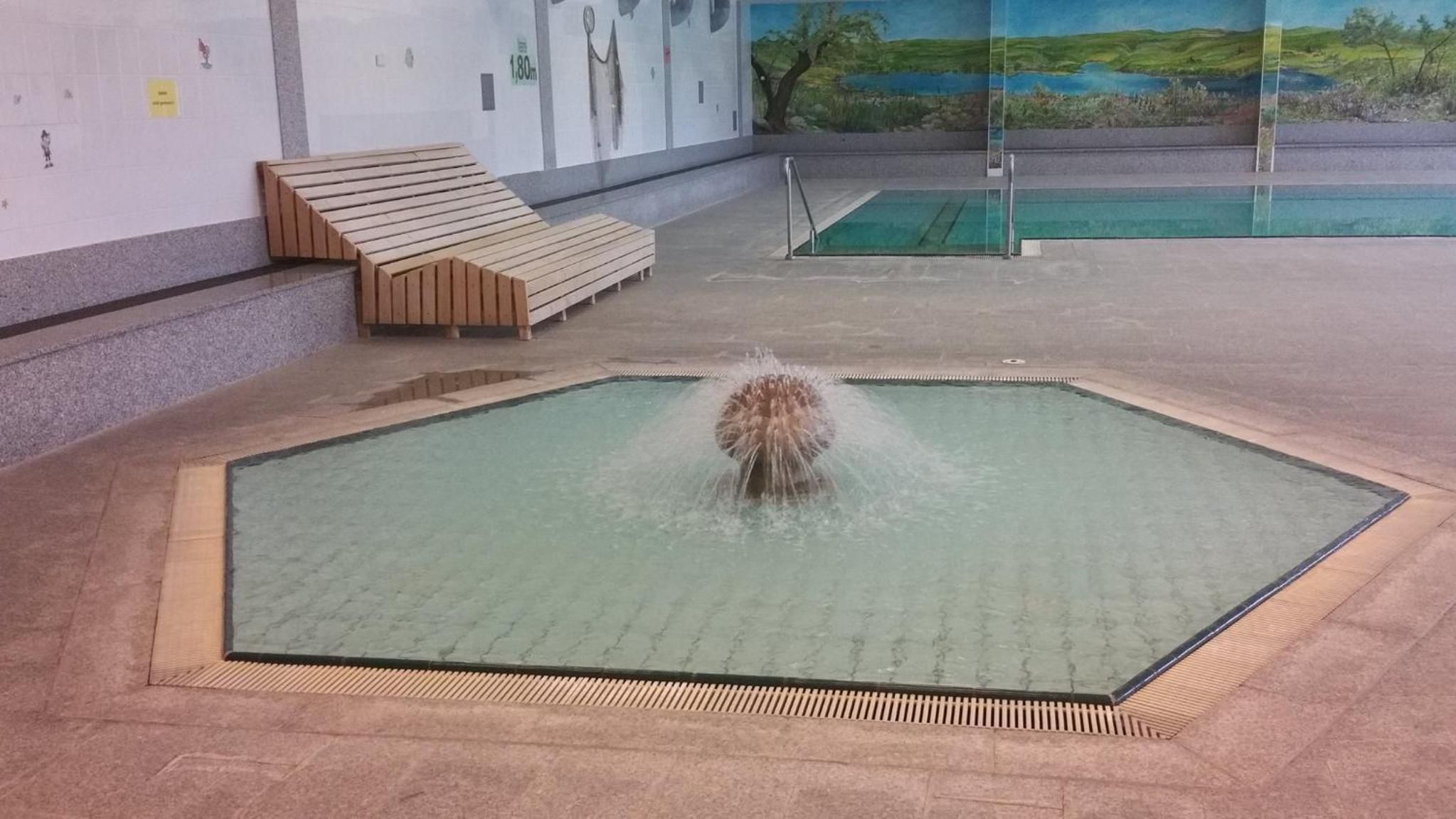 A pool with a water feature in the center, surrounded by a tiled area with a bench and a wall with a mural.