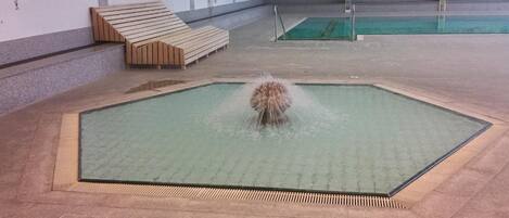 A pool with a water feature in the center, surrounded by a tiled area with a bench and a wall with a mural.