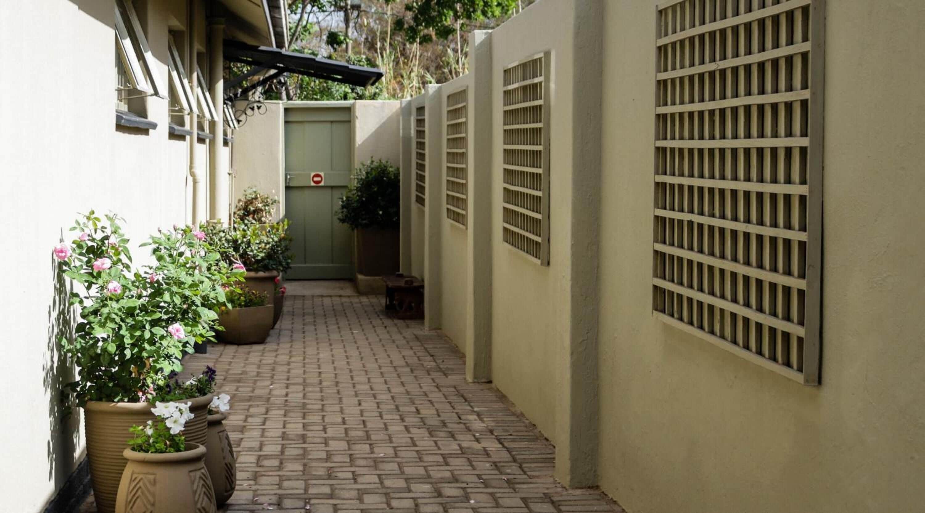 A narrow brick pathway leading to a gated entrance with a green door, flanked by potted plants and a metal gate on the right.
