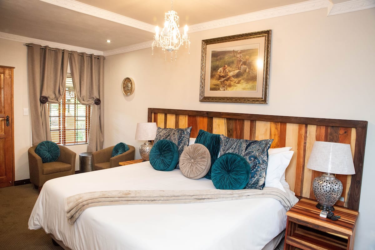 Quarto de hotel com uma cama grande, duas poltronas, uma cabeceira de madeira, um candelabro, um quadro e uma mesa de cabeceira com abajur.
