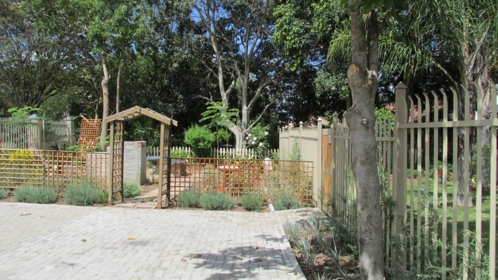 Un chemin pavé mène à une zone délimitée par une grille, où se trouvent une pergola en bois et un jardin clôturé.