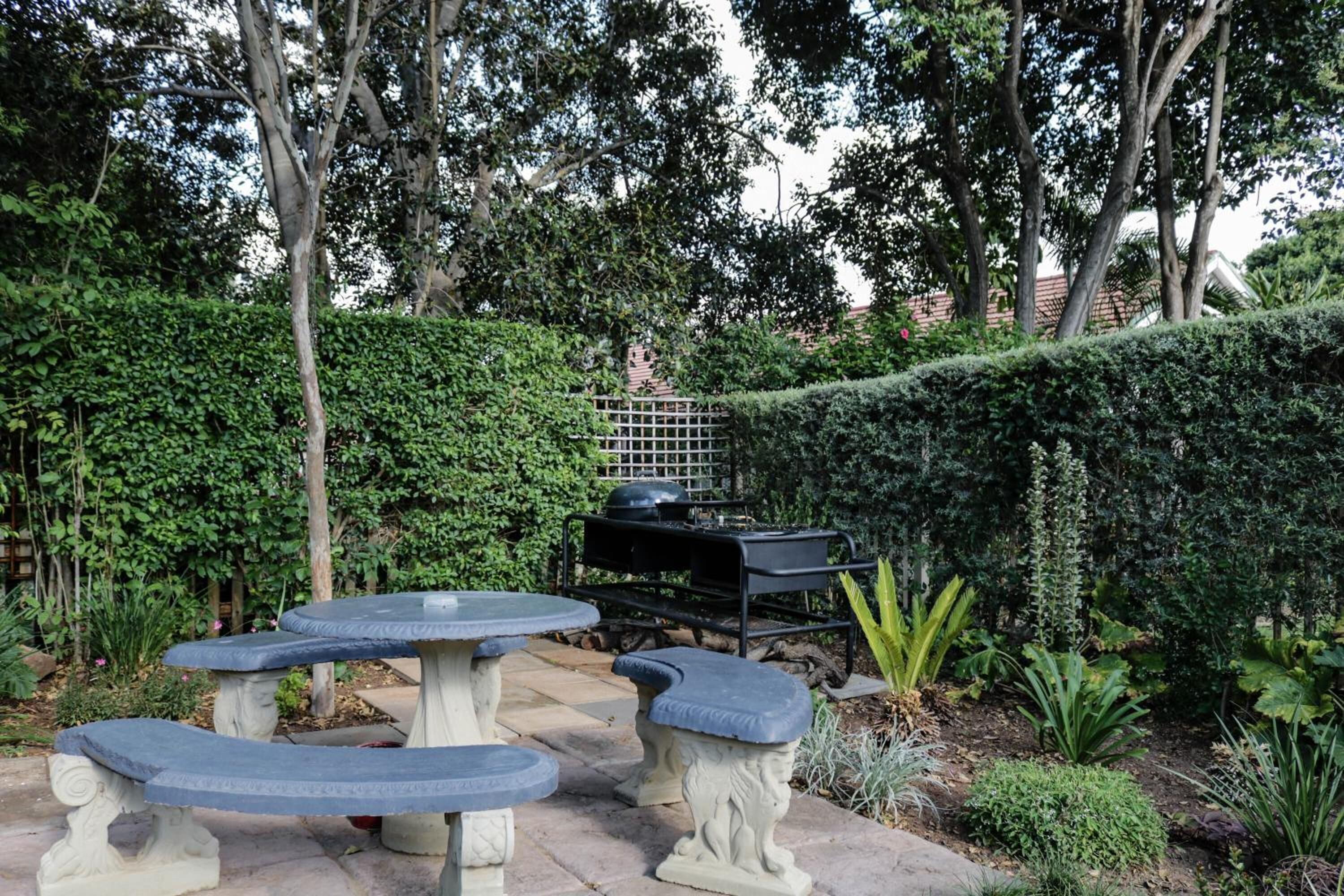 Une terrasse de jardin avec des tables et des bancs en pierre, entourée d’une végétation luxuriante et agrémentée d’un barbecue.