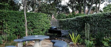 Une terrasse de jardin avec des tables et des bancs en pierre, entourée d’une végétation luxuriante et agrémentée d’un barbecue.