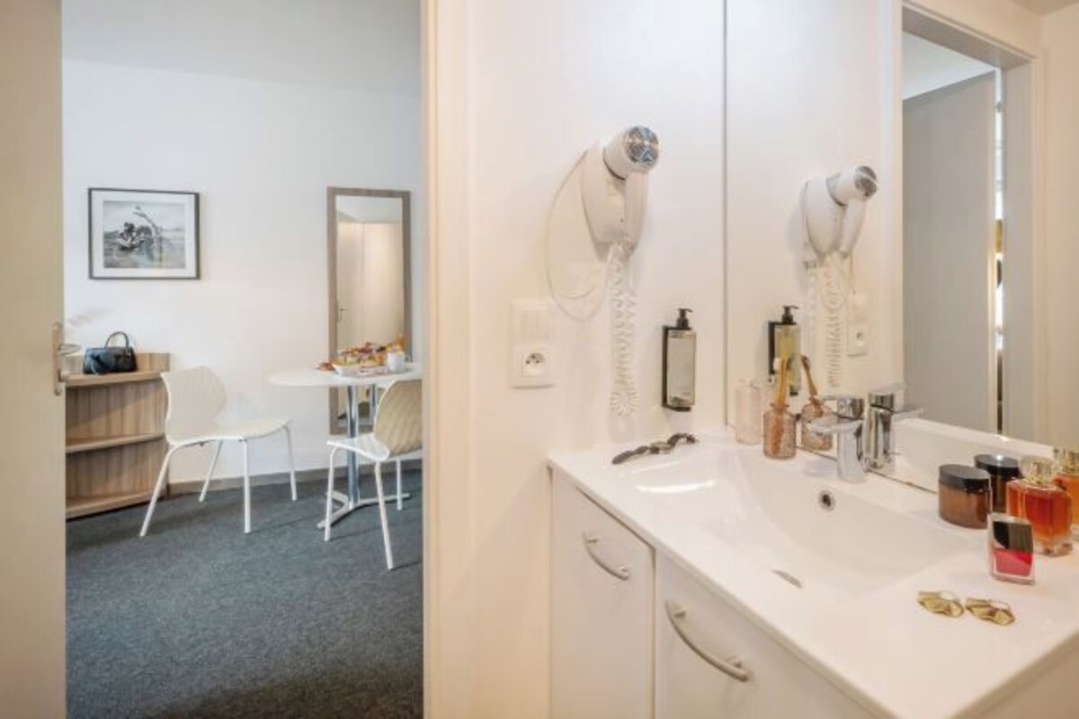 Ett hotellbadrum med ett vitt handfat, hårtork och spegel. Det finns en stol och ett litet bord med toalettartiklar.