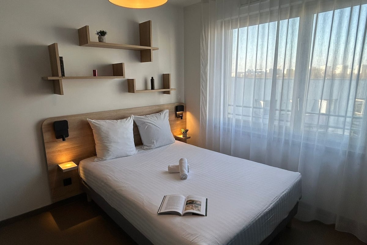 A hotel room with a large bed, bedside lamps, a bedside table, a wall-mounted shelf with items, and a window with sheer curtains.