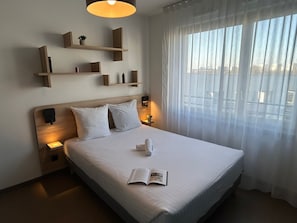 Habitació d'hotel amb un llit gran, abernissadors, una taula de nit, una prestatgeria penjada a la paret amb objectes i una finestra amb cortines translúcides.