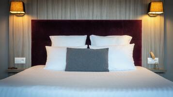 Una cama bien hecha con un cabecero oscuro, dos lámparas de noche y una mesita de noche con un tarro de vidrio.