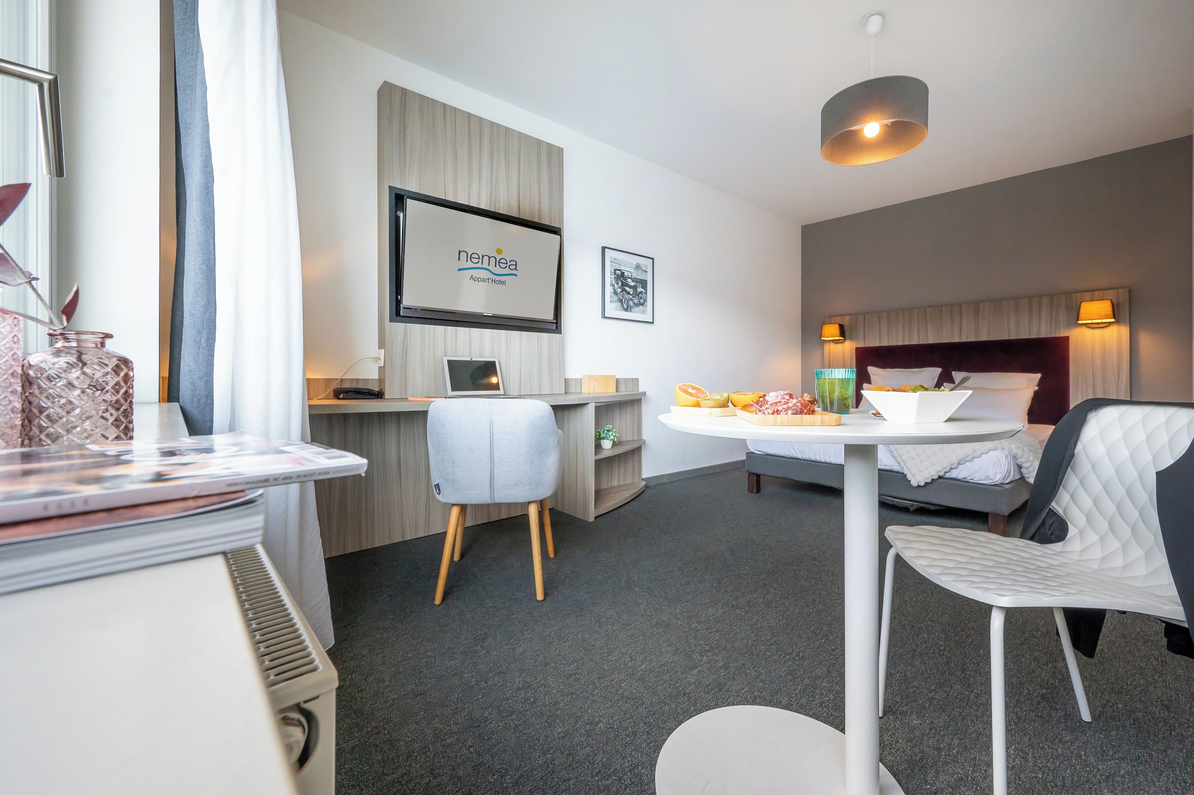 A hotel room with a bed, a desk, a chair, a small table with fruits, a TV, and a picture on the wall.