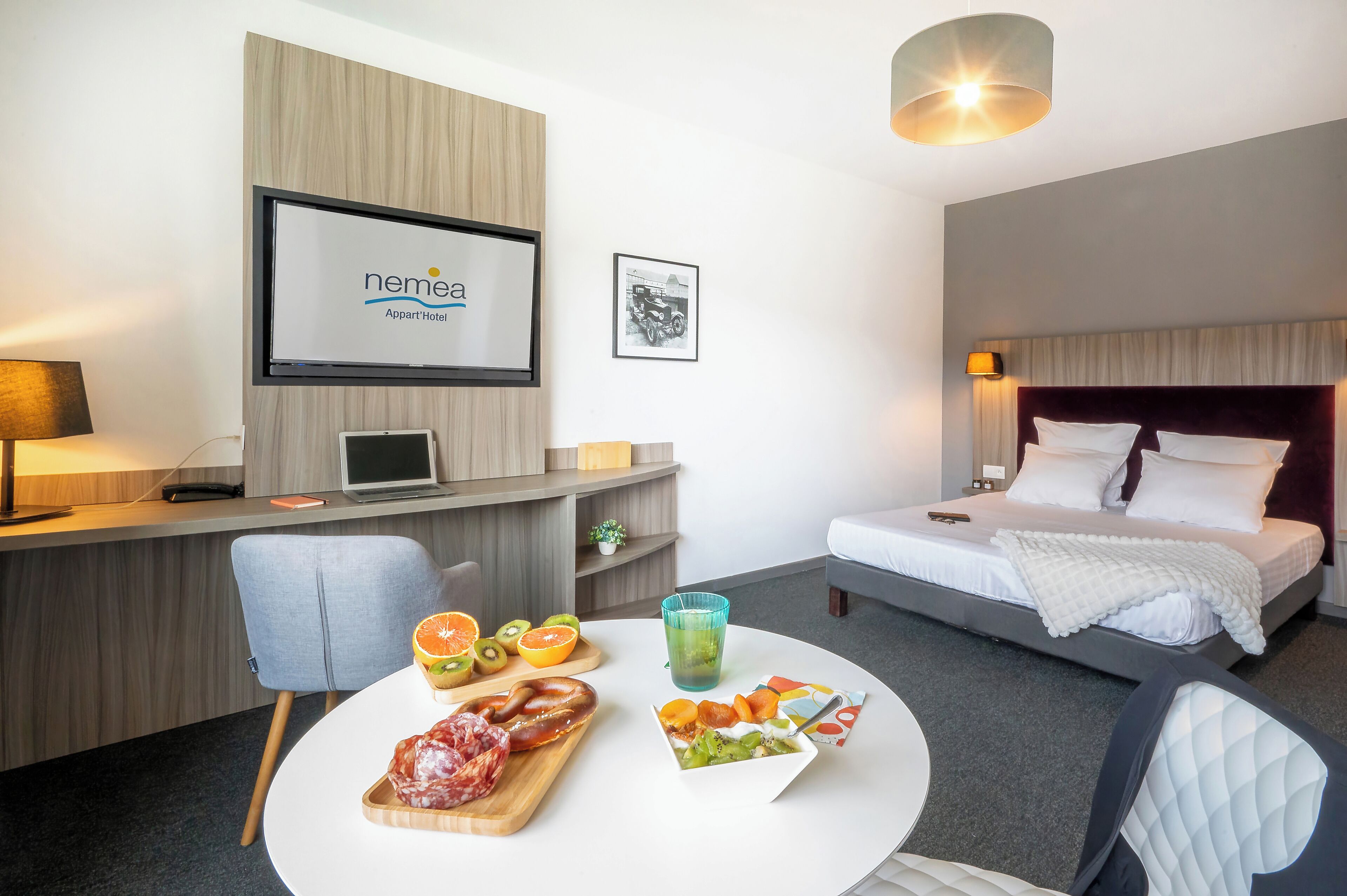 A hotel room with a bed, a desk with a laptop, a TV displaying the hotel name, and a dining table with food and drinks.