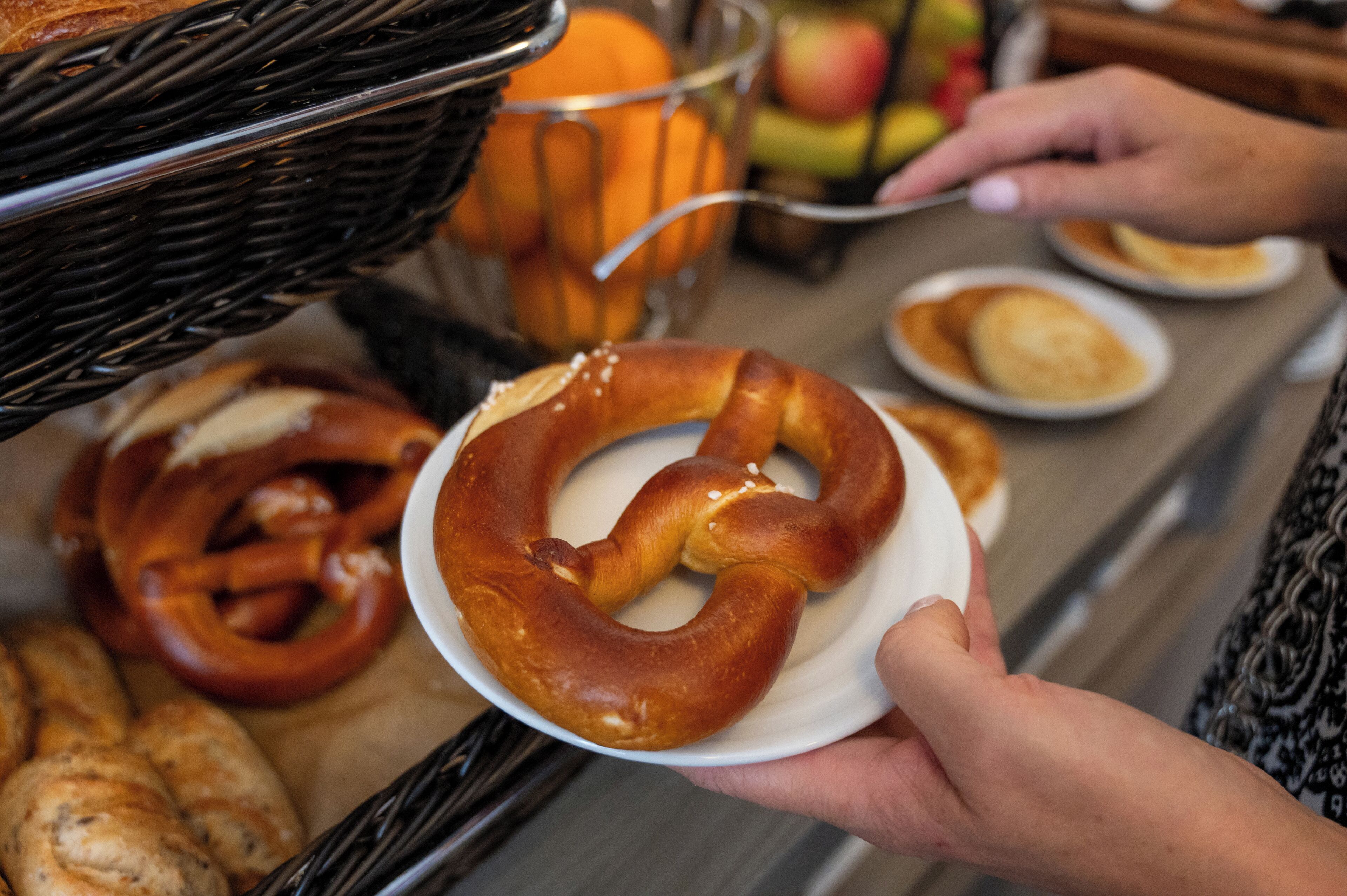 A person holding a pretzel with a basket of pretzels and a bowl of apples in the background.