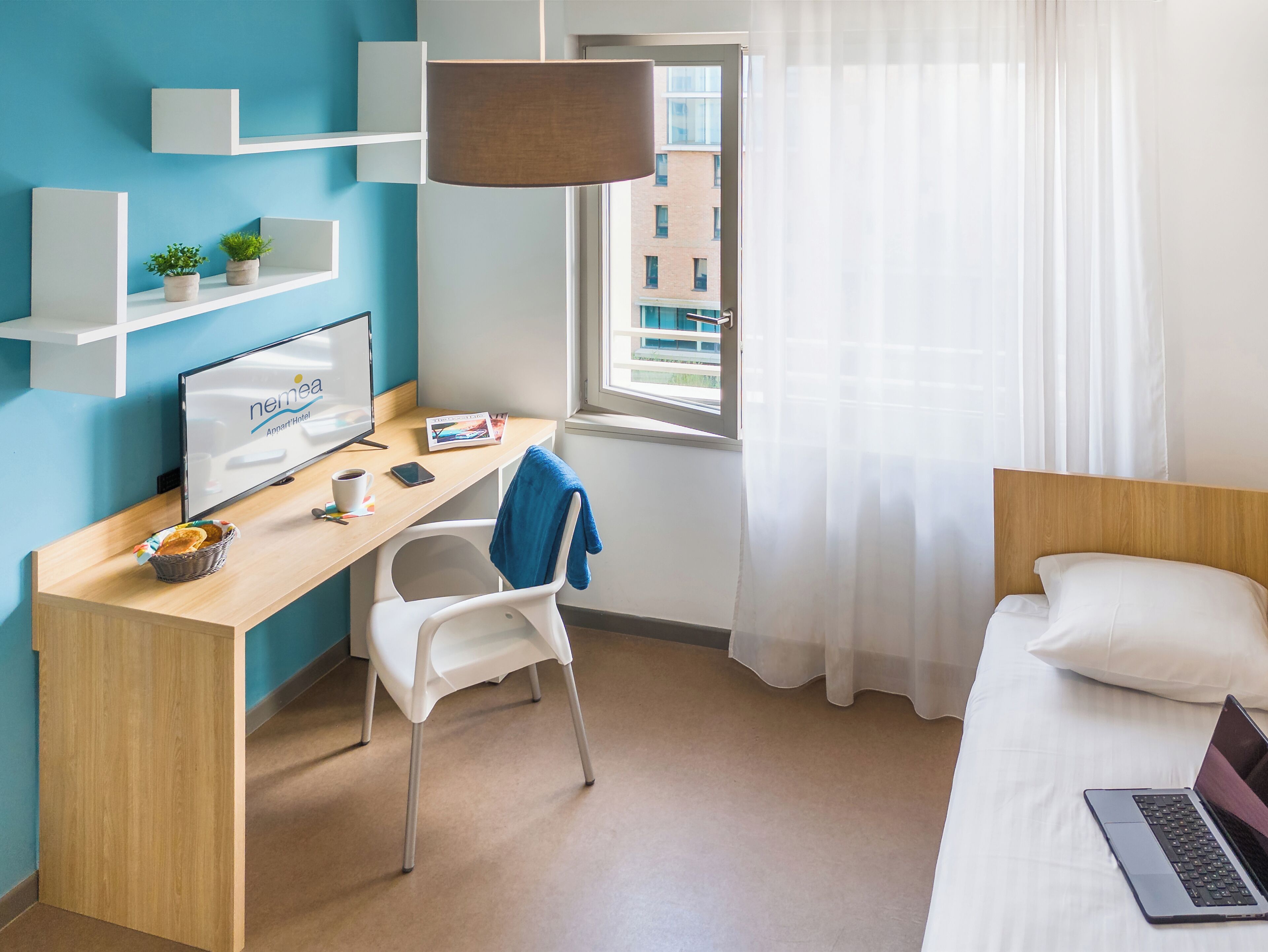 Une chambre d’hôtel avec un bureau en bois, une chaise blanche, un lit avec un oreiller blanc, un ordinateur portable, une fenêtre donnant sur des immeubles et une lampe suspendue.