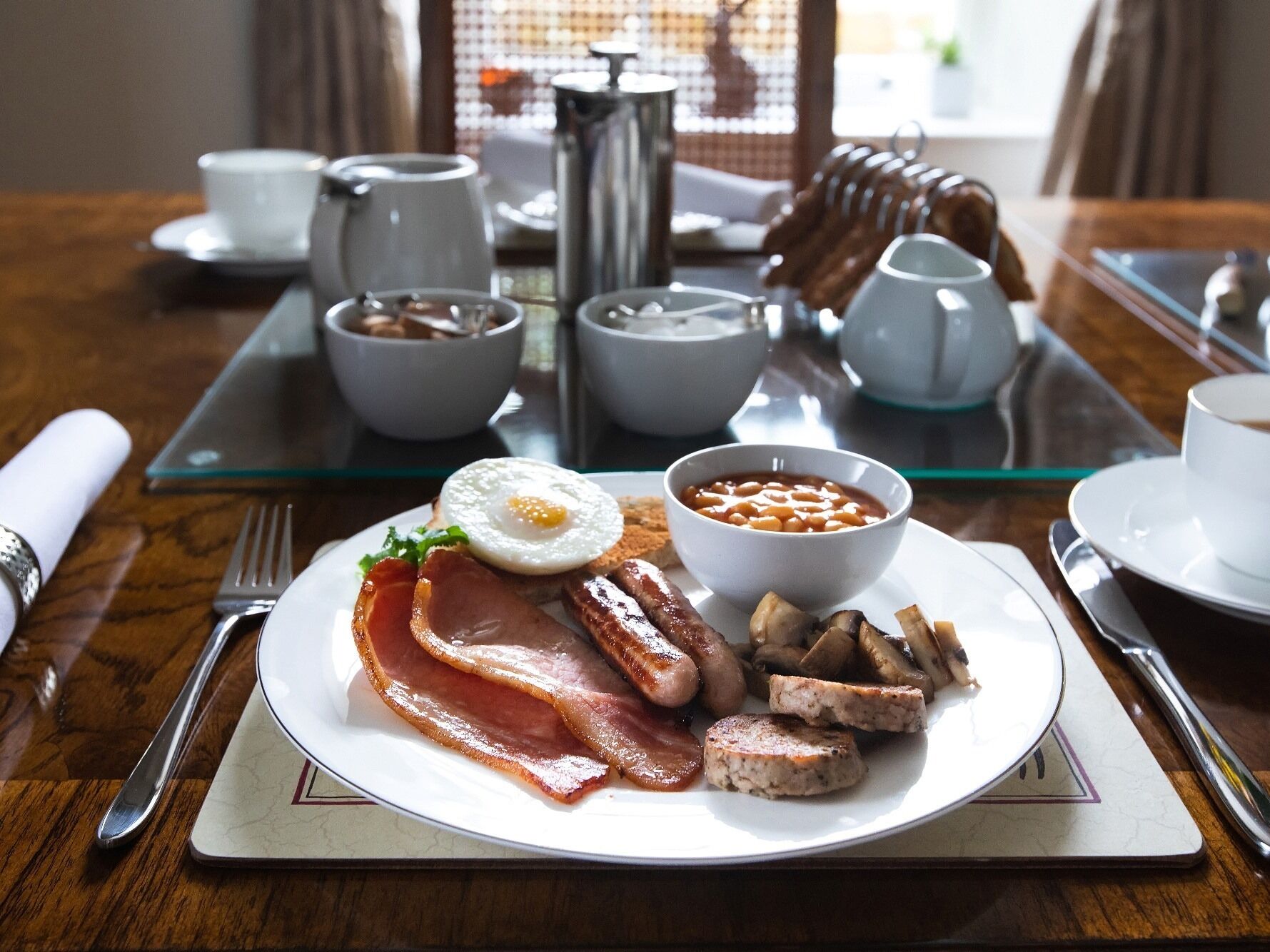 Bandeja de desayuno con tocino, salchichas, champiñones, frijoles y un huevo en un plato, acompañada por una taza de café y una termo.