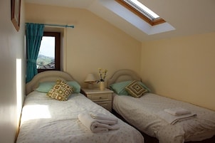 Un dormitorio con dos camas, un tragaluz, una ventana con cortinas, una mesita de noche con una lámpara y un florero con flores.