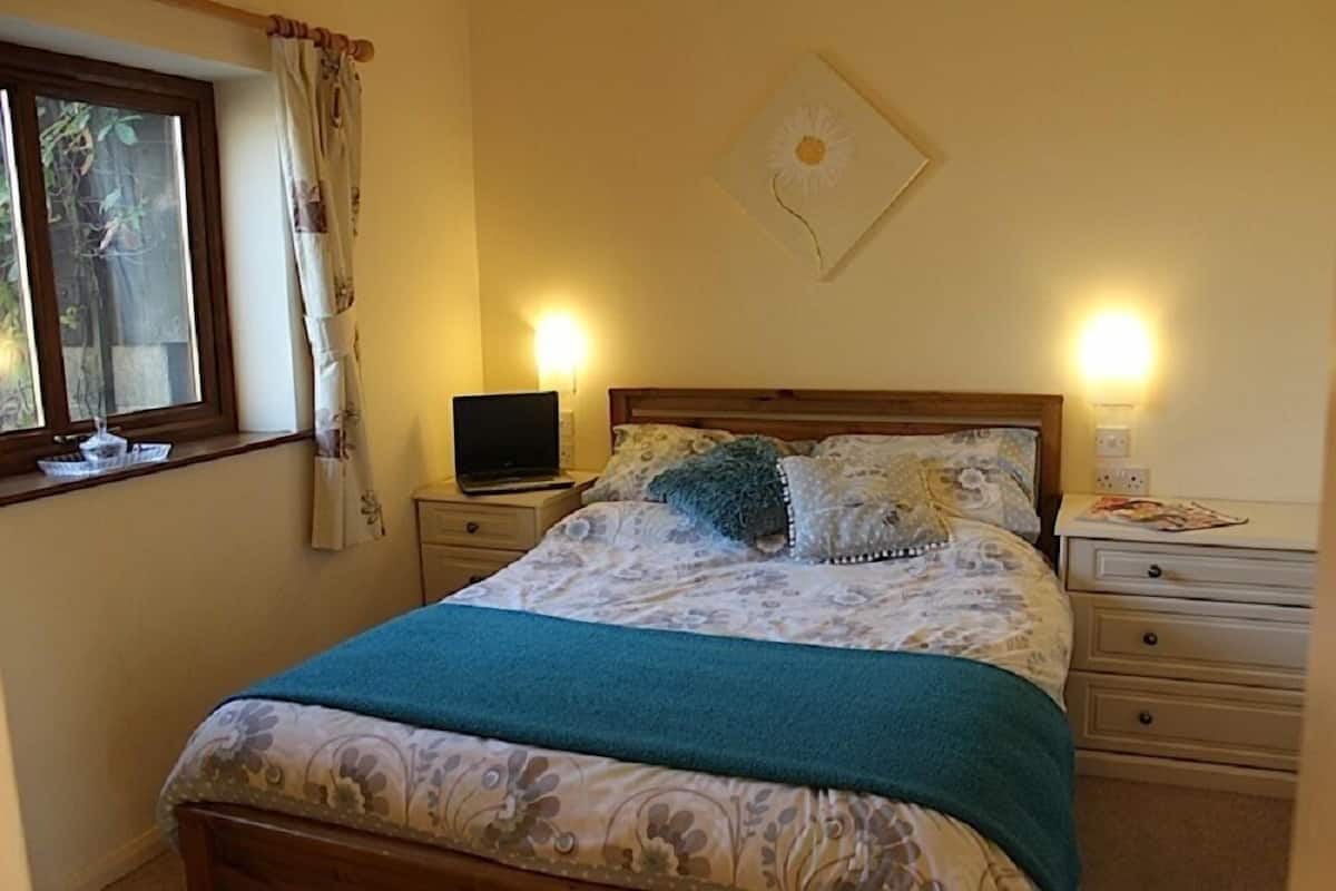 A bedroom with a bed, a nightstand, a laptop, a lamp, and a window with curtains.