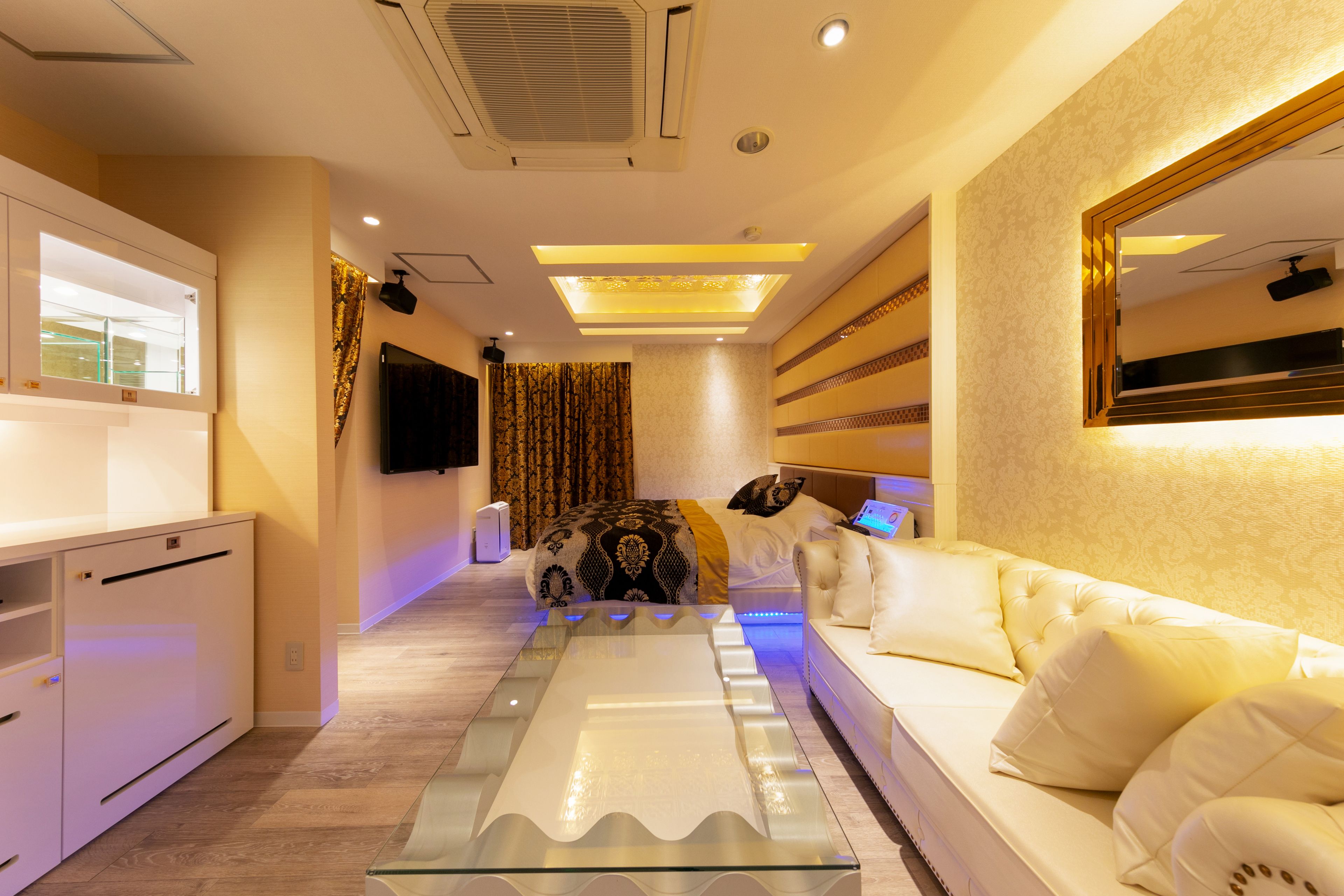 A modern hotel room with a glass coffee table, a white sofa, a flat-screen TV, and a large mirror.
