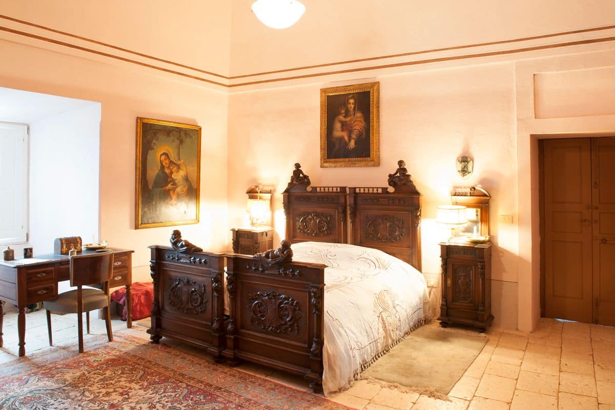 Une chambre à coucher de style traditionnel, avec un grand lit, un bureau et deux chaises. On y trouve deux lampes fixées au mur et un tableau au-dessus du lit.