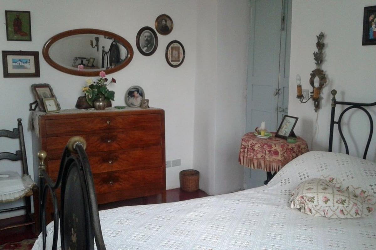 Une chambre avec une commode en bois, un lit recouvert d’une couvre-lit à motifs floraux, une petite table de chevet avec une lampe et un miroir accroché au mur.