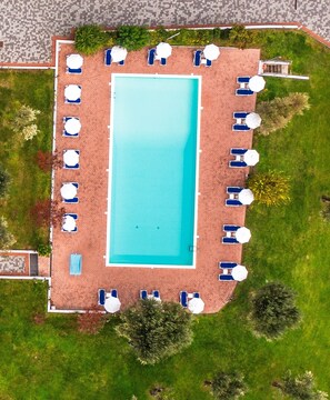 An aerial view of a rectangular swimming pool with a tiled deck surrounding it, equipped with multiple lounge chairs and umbrellas. - Hotel Villa Paradiso Suite  (Moniga del Garda)
