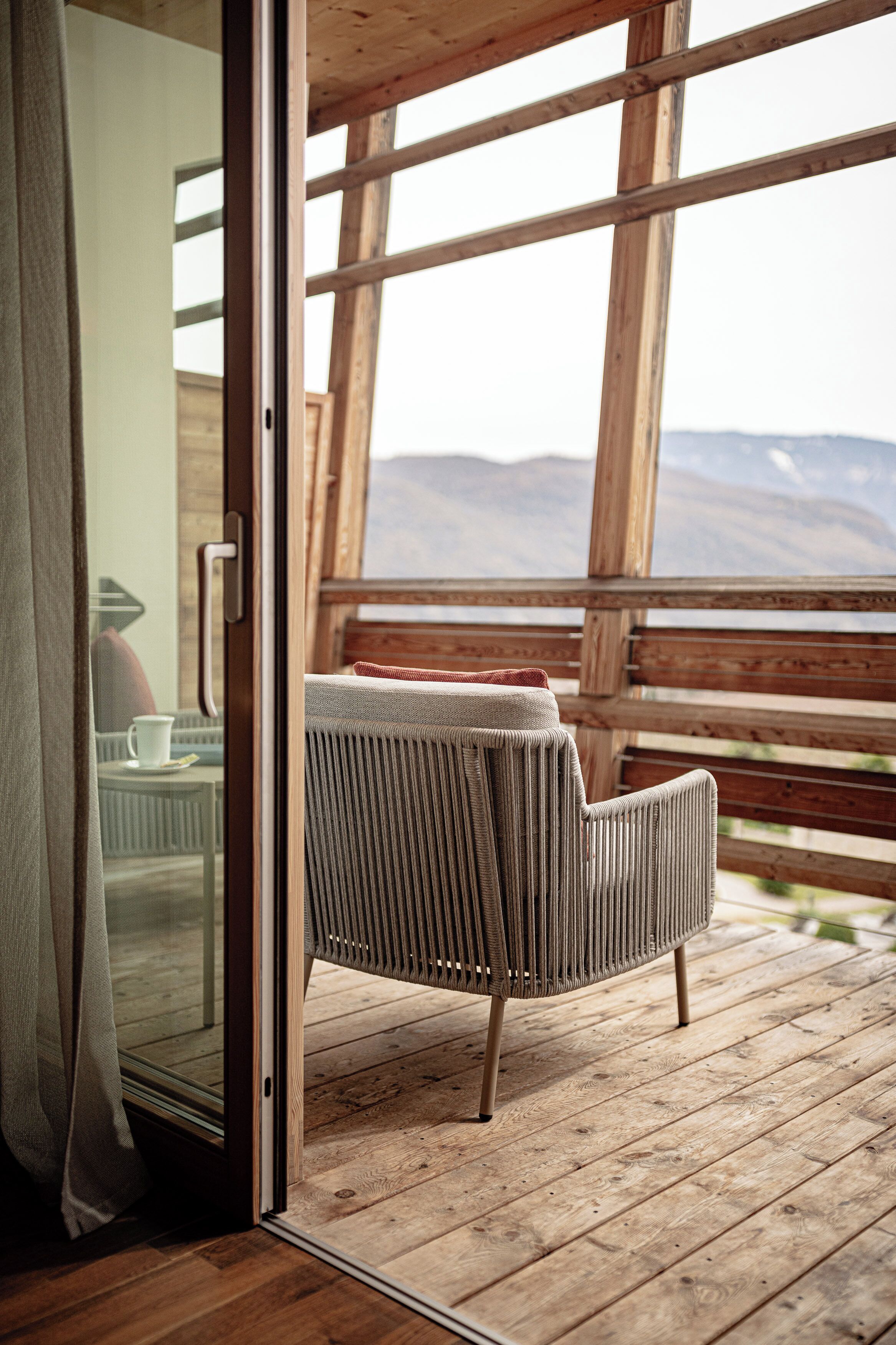 A wooden deck with a wicker chair and a glass door leading to a room with a view of mountains.